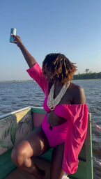 Person in a bright pink cutout swimsuit with billowy sleeves and layered white bead necklace, short dreadlocks, raising a can and enjoying a sunny boat ride on a lake with a distant shoreline.