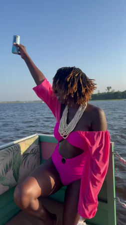 Person in a bright pink cutout swimsuit with billowy sleeves and layered white bead necklace, short dreadlocks, raising a can and enjoying a sunny boat ride on a lake with a distant shoreline.