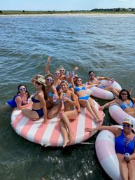 Cheerful group of women in blue swimsuits lounging on a pink-and-white striped inflatable platform and donut floats in coastal bay waters, wearing heart-shaped sunglasses and holding drinks on a sunny summer day.