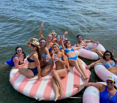 Cheerful group of women in blue swimsuits lounging on a pink-and-white striped inflatable platform and donut floats in coastal bay waters, wearing heart-shaped sunglasses and holding drinks on a sunny summer day.