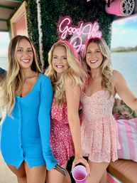Three smiling women in bright summer dresses posing on a boat at the waterfront with a neon "Good Vibes" sign behind them and a pink party cup in hand.