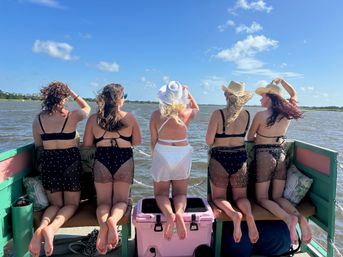 Five friends in bikinis and sheer cover-ups kneeling on the back of a small boat, looking out over a sunny coastal bay with blue sky and marshy shoreline; two wear straw hats and one a white sun hat — summer boat trip vibe.