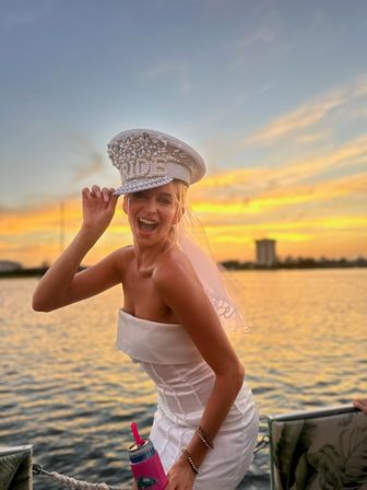 Playful bride in a strapless white dress tipping a jeweled captain’s hat with veil on a boat at golden sunset over a calm waterfront, holding a pink can cooler.