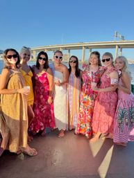 Eight women in colorful summer dresses and sunglasses smiling on a sunny boat cruise by a waterfront bridge, holding drinks and posing for a cheerful group photo.