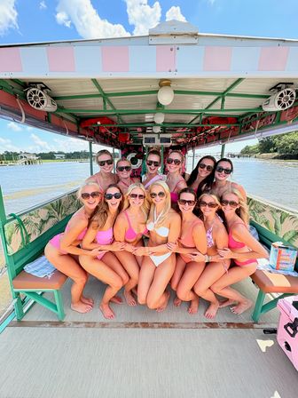 Smiling group of women in pink swimsuits posing together on a colorful covered pontoon boat during a sunny girls' boat party on a coastal river with shoreline and blue sky visible