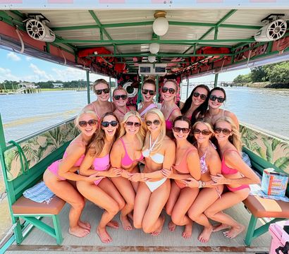 Smiling group of women in pink swimsuits posing together on a colorful covered pontoon boat during a sunny girls' boat party on a coastal river with shoreline and blue sky visible