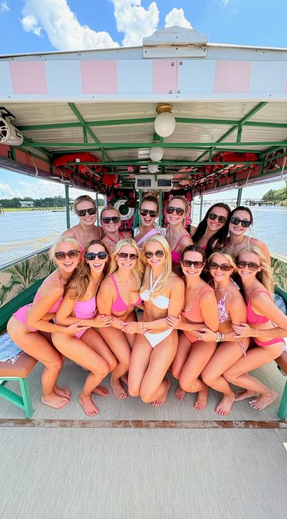 Smiling group of women in pink swimsuits posing together on a colorful covered pontoon boat during a sunny girls' boat party on a coastal river with shoreline and blue sky visible