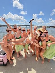 Group of friends in swimsuits lifting a smiling woman on a sunny boat party, holding drinks and a microphone with a bridge and bright blue sky over the water in the background.