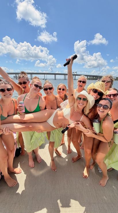 Group of friends in swimsuits lifting a smiling woman on a sunny boat party, holding drinks and a microphone with a bridge and bright blue sky over the water in the background.