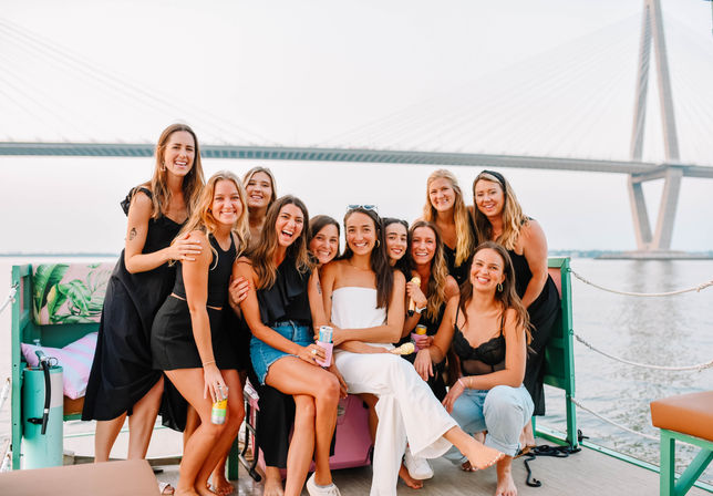 Group of smiling women on a colorful boat by a cable-stayed bridge at the waterfront, casual summer outfits, drinks and a relaxed sunset party vibe.