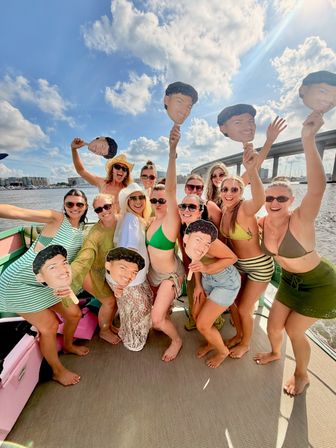 Women in swimsuits on a sunny boat party holding oversized cutout-face paddles, posing by a harbor with a bridge and marina in the background.
