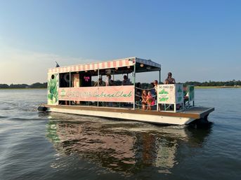 Pink-striped cabana-style pontoon boat with passengers enjoying a sunset cruise on calm coastal South Carolina waters