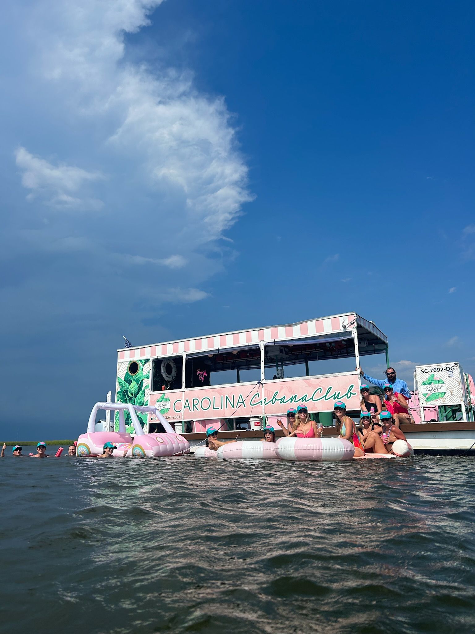 Fun summer scene: group of people on pink-and-white inflatables beside a pastel pink-striped party boat in shallow coastal waters under a bright blue sky with a dramatic billowing cloud.