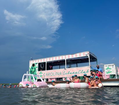 Fun summer scene: group of people on pink-and-white inflatables beside a pastel pink-striped party boat in shallow coastal waters under a bright blue sky with a dramatic billowing cloud.