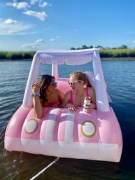 Two friends wearing sunglasses, one with a white veil, lounging on a pink inflatable float-car with a fringe canopy, sipping canned drinks on a calm lake under a bright blue summer sky.