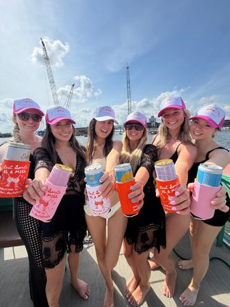 Six friends wearing matching pink-and-white caps and black swim cover-ups toast canned drinks in colorful koozies aboard a boat on a sunny coastal harbor with cranes and blue sky.