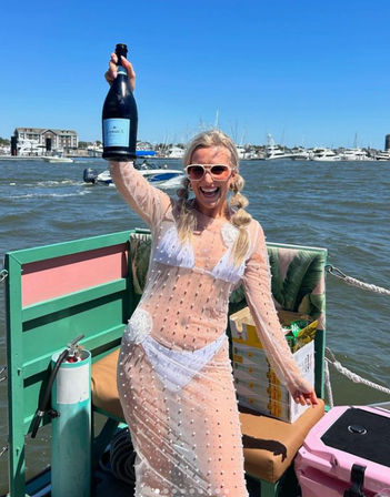 Smiling woman in white bikini and sheer beaded cover-up holding a champagne bottle aboard a colorful boat at a sunny coastal marina with yachts and blue sky