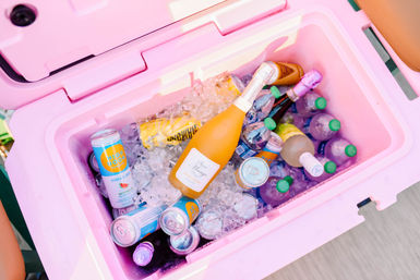 Bright pink portable cooler filled with crushed ice, an orange sparkling wine bottle, assorted canned seltzers and colorful bottled drinks with green caps — vibrant summer party cooler