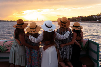 Bachelorette boat at sunset: five women arm-in-arm on a waterfront cruise wearing straw hats—four labeled 'squad' and one white hat labeled 'bride'—watching sailboats and an orange-glow harbor sunset.
