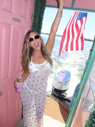 Woman in sunglasses and white crochet cover-up cheering on a pink-decorated boat, holding a canned drink, American flag and outboard motor over a sunny lake