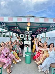 Cheerful group of women in colorful summer dresses raising drinks on a pastel-striped party boat cruising a coastal harbor with a bridge and blue cloudy sky in the background