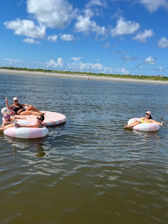 Group of friends lounging on pink-striped inflatable tubes and a round float in calm coastal waters near a sandy beach under a bright blue sky with fluffy clouds