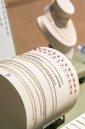 Close-up jewelry display of delicate gold chain bracelets and beaded gemstone wristlets on a cream leather roll, pink and blue stones with a blurred necklace bust in the background — retail jewelry showcase.