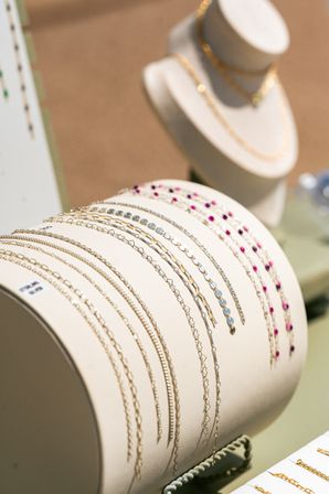 Close-up jewelry display of delicate gold chain bracelets and beaded gemstone wristlets on a cream leather roll, pink and blue stones with a blurred necklace bust in the background — retail jewelry showcase.