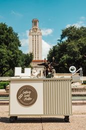 Cream rolling jewelry cart with necklace displays and magnifying lamp at an outdoor pop-up, fountain and tall clock tower framed by trees in Austin, Texas on a sunny day