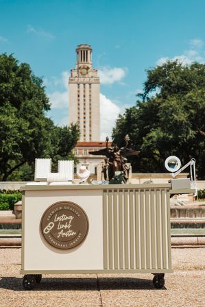 Cream rolling jewelry cart with necklace displays and magnifying lamp at an outdoor pop-up, fountain and tall clock tower framed by trees in Austin, Texas on a sunny day