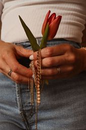 Close-up of hands holding a red tulip and tangled gold chains over blue jeans and a white ribbed top, fingers adorned with stackable rings.