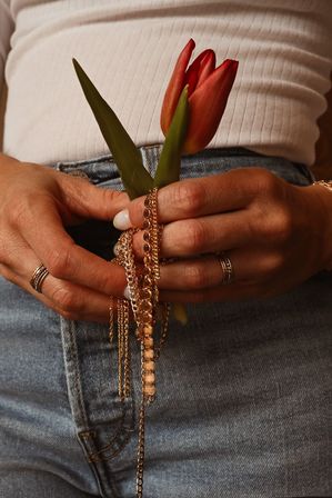Close-up of hands holding a red tulip and tangled gold chains over blue jeans and a white ribbed top, fingers adorned with stackable rings.