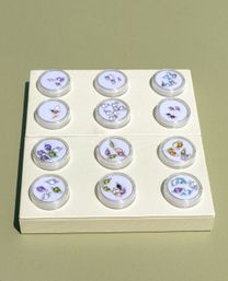Flat-lay jewelry display: cream board with twelve clear round cases in a 3x4 grid, each holding colorful teardrop and marquise gemstone charms (purple, green, blue, amber) on a muted olive-green background.