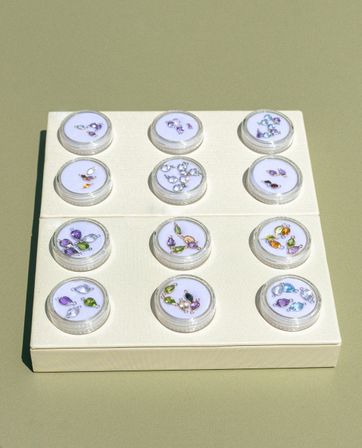 Flat-lay jewelry display: cream board with twelve clear round cases in a 3x4 grid, each holding colorful teardrop and marquise gemstone charms (purple, green, blue, amber) on a muted olive-green background.