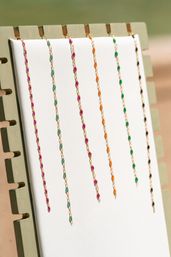 Sunlit jewelry display of delicate gold chain necklaces with colorful bead accents—pink, teal, orange, green and black—hanging vertically on a white board at an outdoor market.
