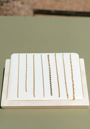 Product photo of multiple delicate gold chains laid vertically on a white padded display atop a sage-green tabletop — minimalist jewelry styling for necklaces and bracelets.