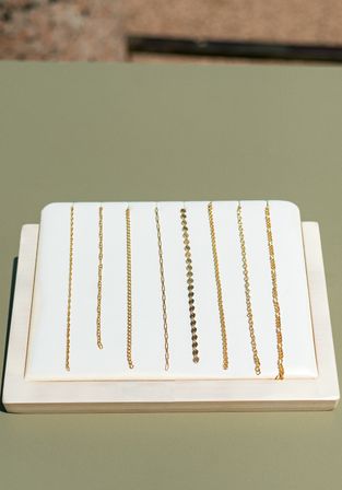 Product photo of multiple delicate gold chains laid vertically on a white padded display atop a sage-green tabletop — minimalist jewelry styling for necklaces and bracelets.