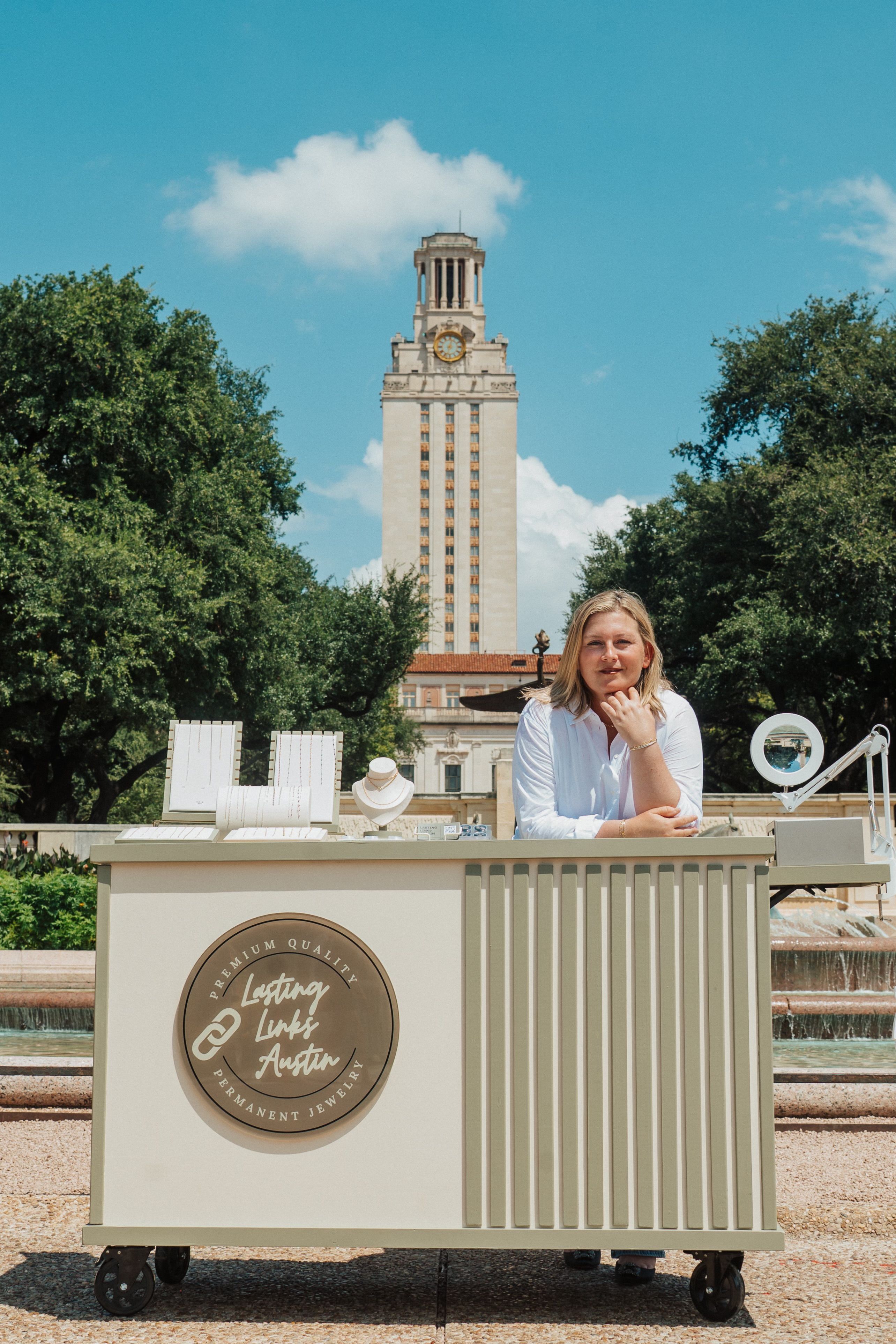 Outdoor jewelry pop-up cart with necklace displays and a vendor leaning on the counter, fountain and a tall clock tower framed by trees under a bright blue sky