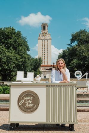 Outdoor jewelry pop-up cart with necklace displays and a vendor leaning on the counter, fountain and a tall clock tower framed by trees under a bright blue sky