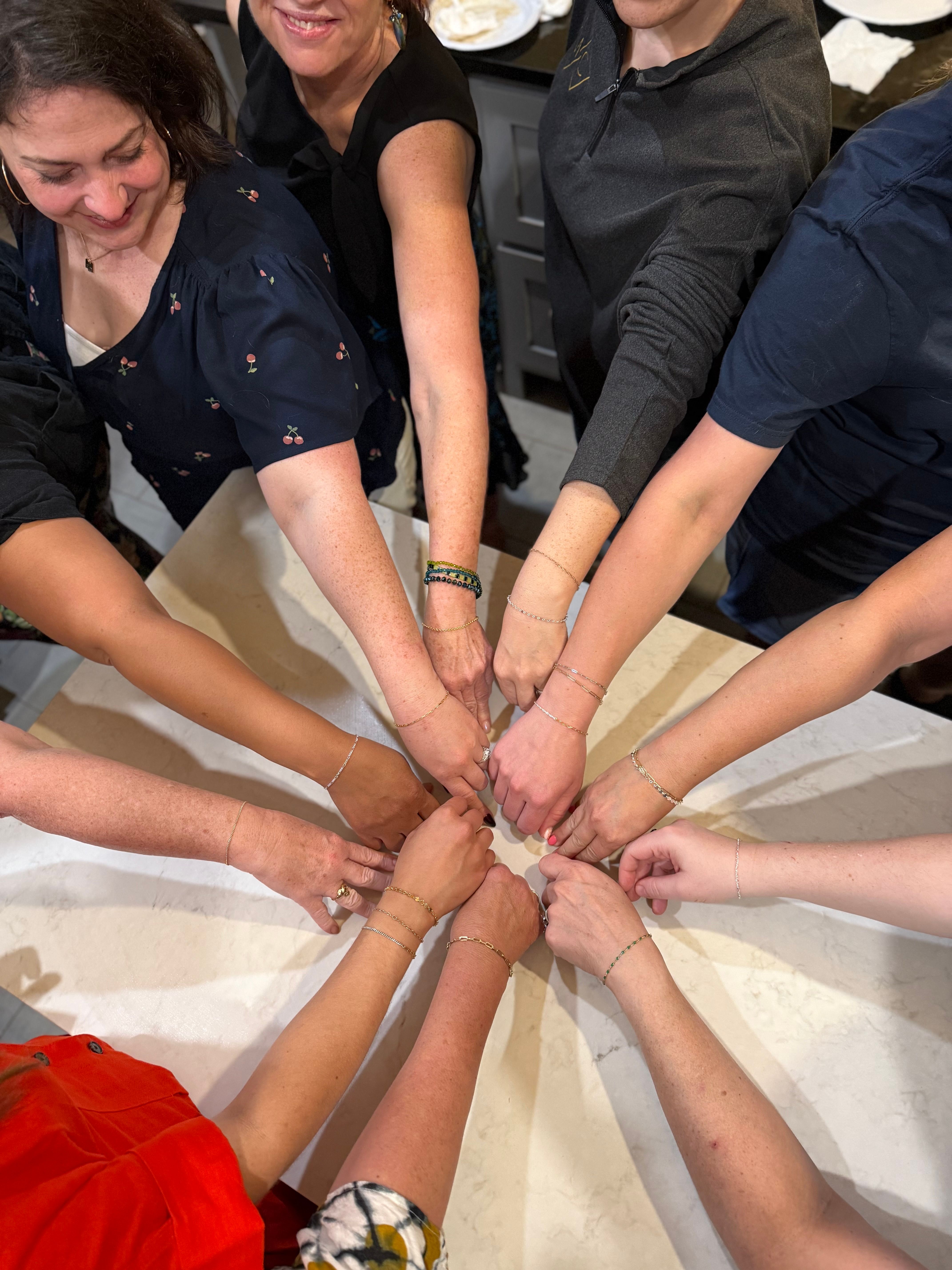 Overhead shot of a group of people around a kitchen island, arms extended inward to form a circle and display matching delicate bracelets on their wrists — cheerful bracelet-gifting moment.