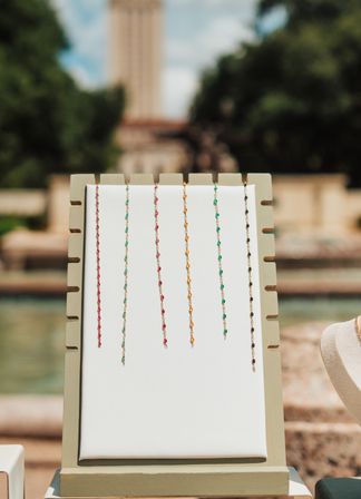 Sunlit outdoor jewelry display board with seven delicate beaded chains in pink, turquoise, gold, green and black hanging against a blurred park fountain backdrop.