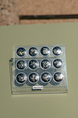 Clear acrylic display tray with twelve round compartments holding small gold-filled alphabet and symbol charm pendants, labeled 'GOLD FILLED', photographed on a pale olive-green tabletop in sunlight.
