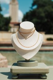Sunlit outdoor necklace display on a cream fabric bust featuring layered delicate gold chains with a small green gemstone, blurred fountain and trees in the background.