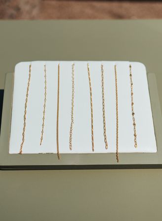 Close-up of delicate gold chains neatly lined vertically on a white jewelry display tray over an olive-green surface, minimalist necklace showcase.