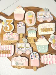 Assorted pastel sugar cookies decorated as ice cream cones, cupcakes, popsicles, an ice cream truck and a number 3, arranged on a round wooden board for a sweet third-birthday celebration.