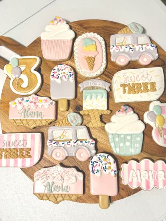 Assorted pastel sugar cookies decorated as ice cream cones, cupcakes, popsicles, an ice cream truck and a number 3, arranged on a round wooden board for a sweet third-birthday celebration.