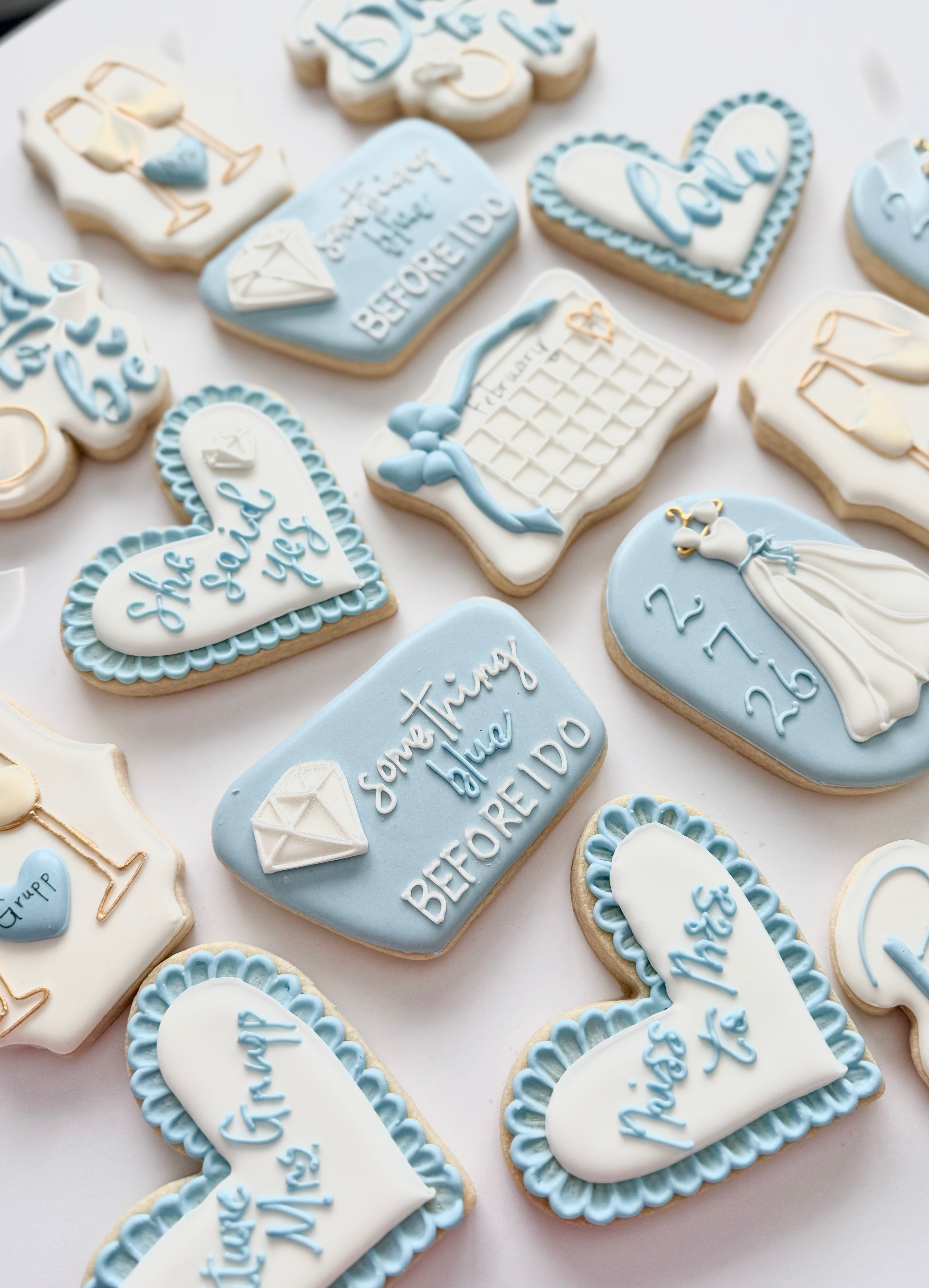 Assorted pastel blue and white iced sugar cookies styled for a bridal shower or engagement—heart shapes, diamond ring, wedding dress and calendar designs with 'something blue' messages on a white background.