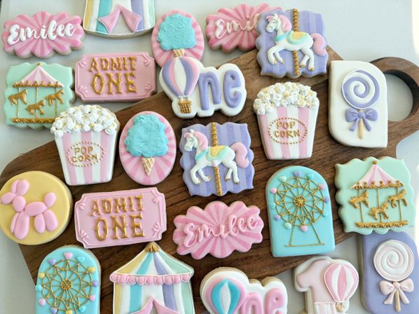 Pastel carnival-themed iced sugar cookies arranged on a wooden board, featuring popcorn tubs, carousel horses, ferris wheel, hot-air balloons, cotton candy, lollipops, ‘Admit One’ tickets and a number one for a first birthday party