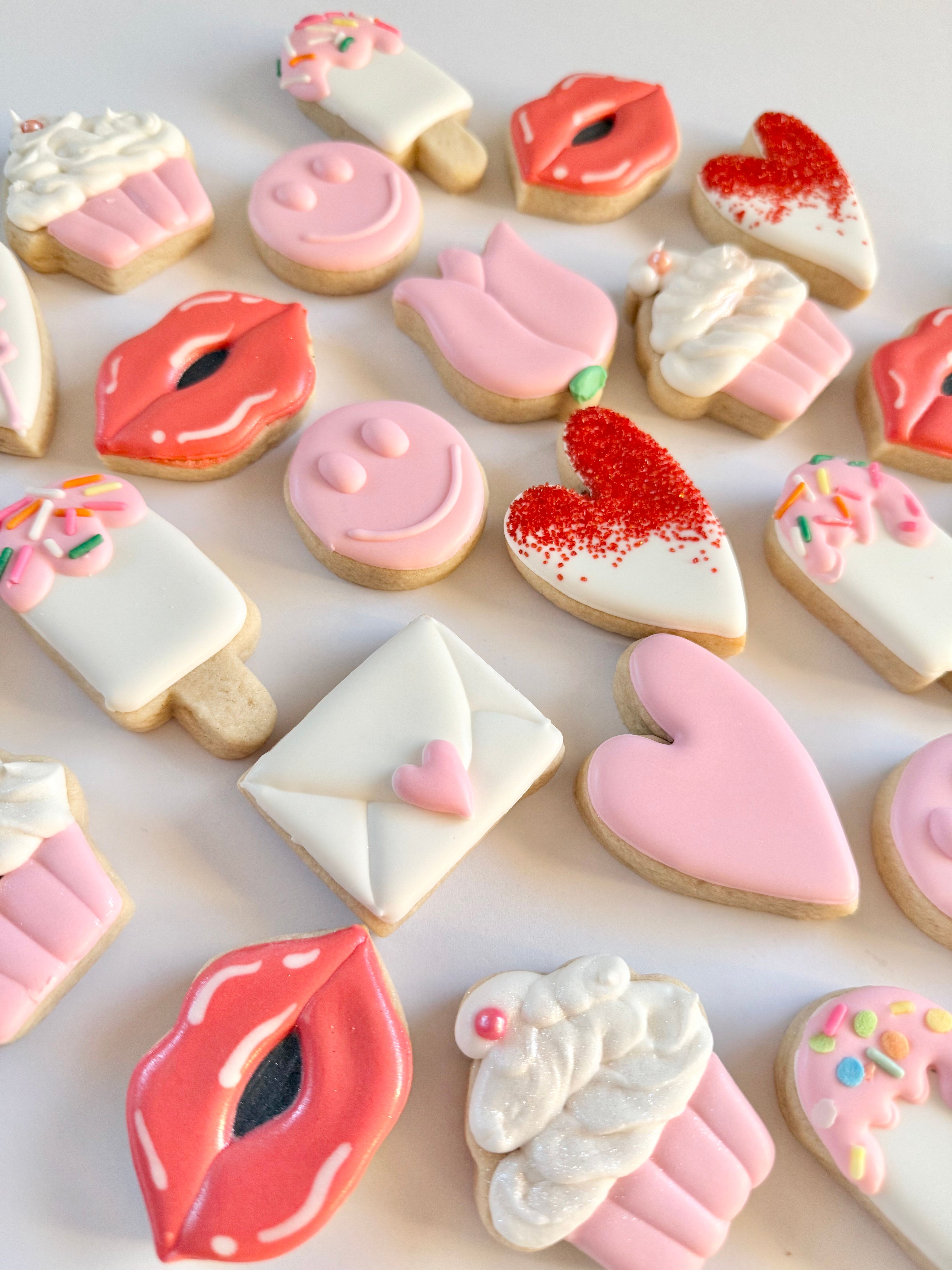 Valentine-themed flatlay of assorted iced sugar cookies in pink, red and white — hearts, lips, cupcakes, popsicles, tulips, smiley faces and love-letter shapes on a white background