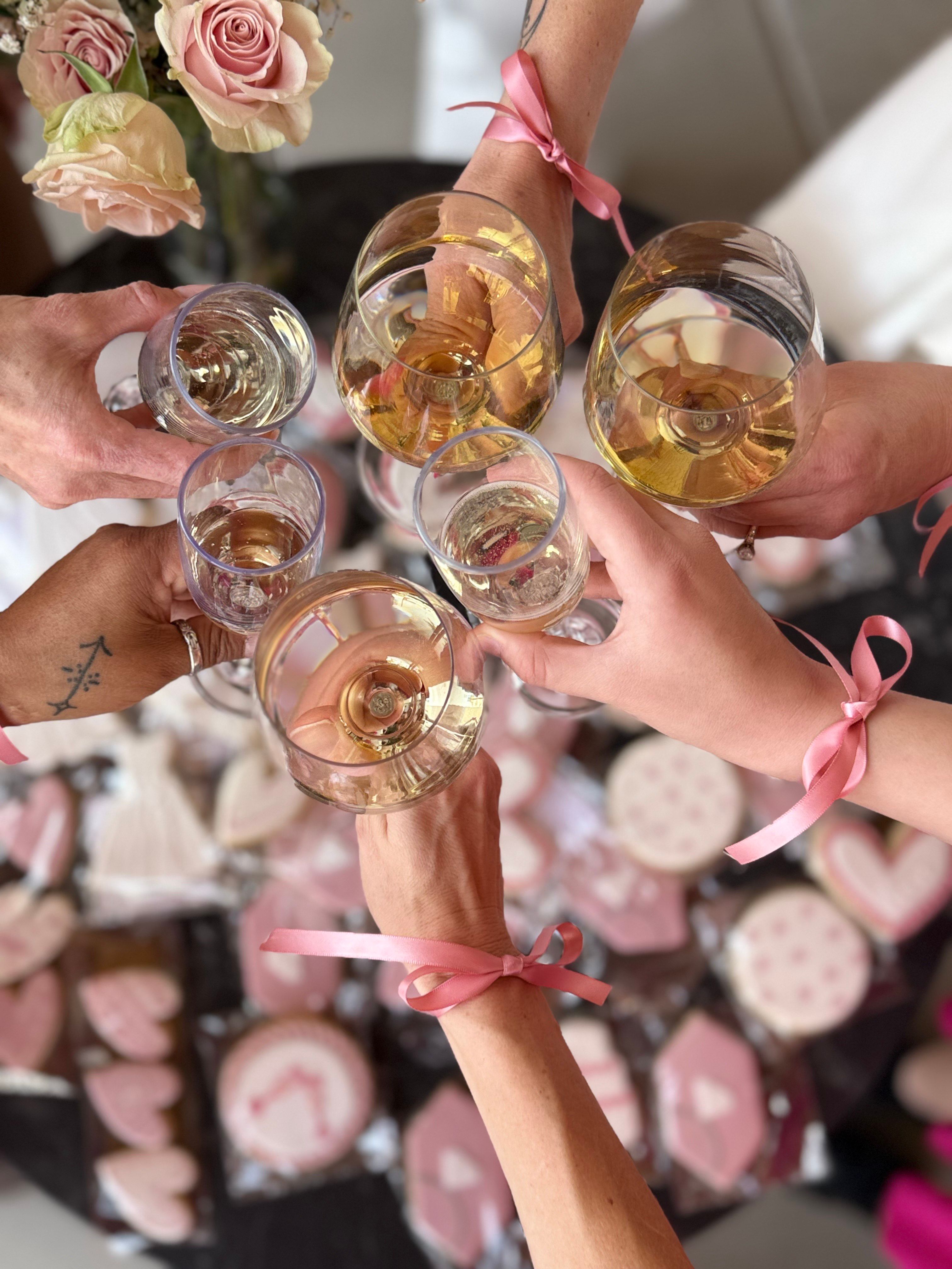 Top-down view of a champagne toast at a bridal shower — hands tied with pink ribbons hold wine and champagne glasses over a table of pink heart-shaped decorated cookies and pale roses